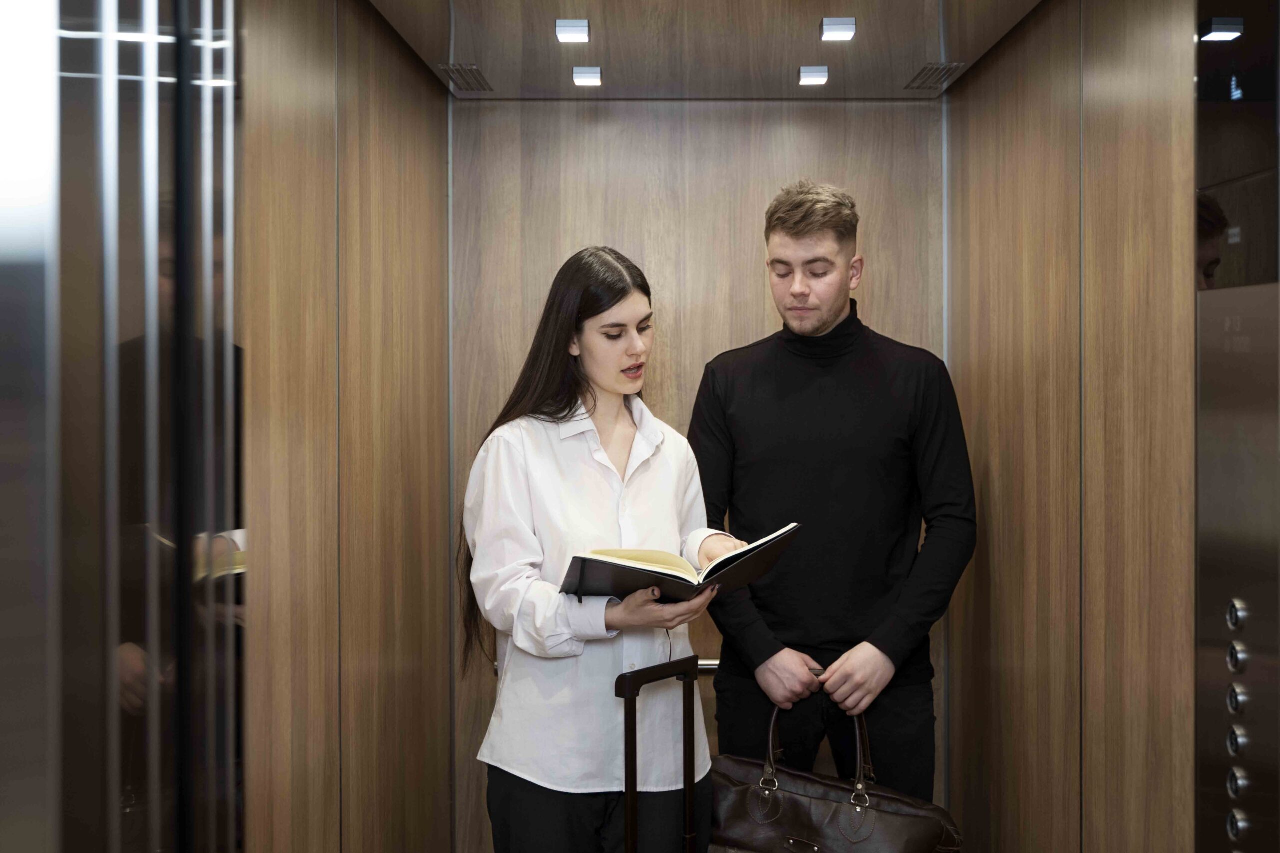 Un jeune couple est dans un ascenseur. La femme tient un livre ouvert et le montre à son ami. Une valise est posée devant eux.